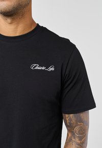 Άνδρας που φορά μαύρο t-shirt με λευκό λογότυπο «Closure Ldn» στο στήθος και ορατό τατουάζ τριαντάφυλλου στον δεξί πήχυ, με φόντο απλό χρώμα.