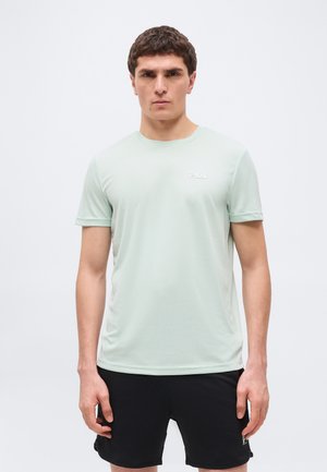 Mand iført lysegrøn FILA kortærmet sportstrøje og sorte shorts, stående mod en ensartet lys baggrund.