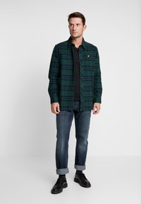Chemise à carreaux verte et marine avec fermeture à boutons, manches longues et logo sur la poitrine, associée à un jean foncé et des chaussures noires.