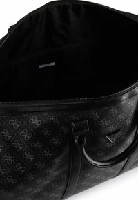 Bolsa tipo duffle negra con superficie texturizada, patrón de logo, cierre de cremallera y dos asas. Interior forrado con etiqueta de marca visible.