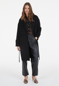 Schwarzer Wollmantel mit großen Revers, strukturierter Form, Knopfverschluss, getragen über einer schwarzen Spitzenbluse. Kombiniert mit schwarzen Lederhosen und Riemchen-Heels.