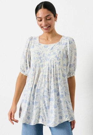 Frau mit dunklen Haaren in einem tiefen Dutt trägt eine weiße Bluse mit blauem Blumenmuster, Puffärmeln, plissiertem Vorderteil und hellblauen Jeans, lächelt und schaut nach unten.