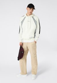 Felpa bianca con dettagli grigi e maniche nere, pantaloni cargo beige, sneaker bianche e una borsa in vita rosso scuro con un logo.