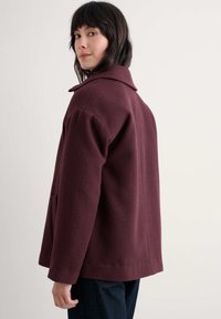 Burgundowa tkana kurtka z wyciętym kołnierzem, bocznymi kieszeniami i luźnym krojem. Teksturowany materiał widoczny na powierzchni.