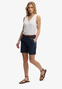 Mujer con top blanco sin mangas y escote en V, pantalones cortos azul marino con cinturón trenzado marrón, y sandalias planas marrones, caminando con una mano en el bolsillo.