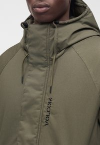 Gros plan d'une personne portant une veste à capuche vert olive avec cordons et un logo "Volcom" noir vertical sur le devant.