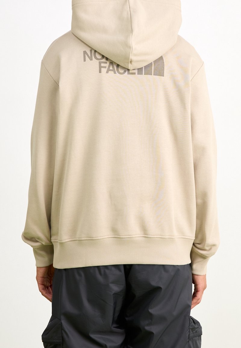 Sweat à capuche beige avec poignets et ourlet côtelés, présentant un logo partiel sombre sur le haut du dos, porté avec un pantalon noir.