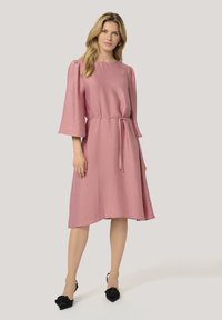 Rosa klänning i knälängd med 3/4 ärmar och midjeband. Lättviktig, texturerad tyg med en lös passform. Bärs med svarta skor som har blomsterdetaljer.