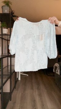 Blusa azul clara, semi-transparente, com mangas bufantes de três quartos, apresentando um padrão geométrico subtil e um decote redondo.