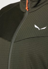 Tmavozelená bunda na zips s vysokým golierom, štruktúrovanou tkaninou a kontrastným bielym logom na hrudi, doplnená oranžovým zipsom.