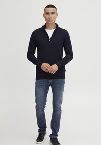 Navy Zip-Hoodie aus Strickstoff, kombiniert mit blauen Jeans und schwarzen Sneakers. Der Pullover hat einen hohen Kragen und gerippte Bündchen.
