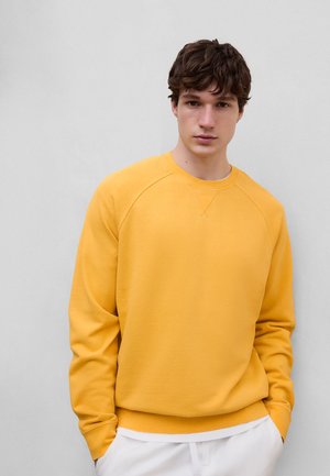 Ung mann med brunt hår iført en okergul genser og hvite bukser står mot en ensfarget lys grå vegg.