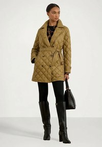 Egy övvel ellátott, steppelt olíva-zöld kabátot viselő nő, fekete leggingsben, térd fölé érő fekete csizmában és fekete kézi táskával, egy fehér háttér előtt áll.