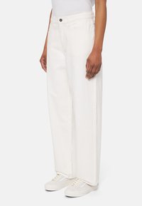 Jeans blancos de pierna ancha de denim, con cintura alta, bolsillos frontales y cierre de botón, combinados con zapatillas de color claro.