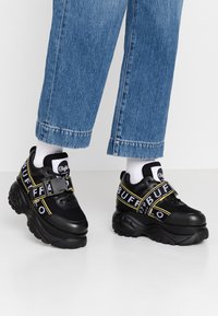 Svarta sneakers med tjock sula, som har meshnät och syntetiskt material. Djärv "BUFF"-logga i vitt och gult på remmarna.