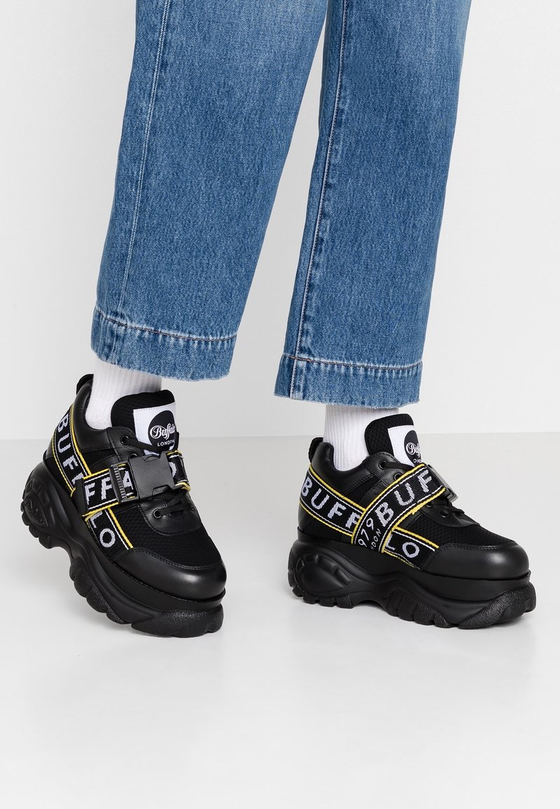 Svarta sneakers med tjock sula, som har meshnät och syntetiskt material. Djärv "BUFF"-logga i vitt och gult på remmarna.