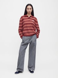 Femme debout, les mains dans les poches, portant une chemise rouge à rayures manches longues, un pantalon gris ample et des baskets bleu marine sur fond blanc.