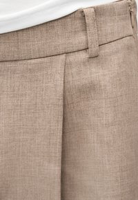 Μπεζ υφασμάτινο παντελόνι με θηλιές για ζώνη και πιέτες, σε συνδυασμό με λευκή μπλούζα που είναι μπατισμένη στη μέση.