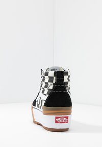 Zapatilla negra y blanca de cuadros en estilo high-top con acentos de gamuza, franjas blancas y una suela de goma que presenta un logo rojo de "Off The Wall".