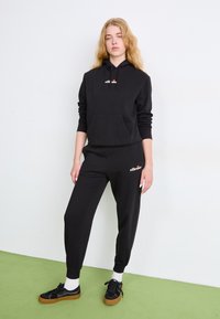 Zwarte hooded sweatshirt en joggers van een zachte stof, met een voorzak en logo-accenten. Model draagt zwarte schoenen met witte sokken.