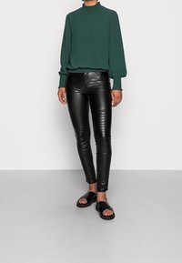 Mörkgrön blus med rynkad halsringning, i kombination med svarta fake läder leggings med ribbade detaljer på låren och svarta platta sandaler.