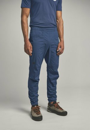 Mann trägt blaue Hosen mit elastischem Bund, ein dunkelblaues T-Shirt und graue Wanderschuhe mit orangen Schnürsenkeln, steht vor einfarbigem Hintergrund.