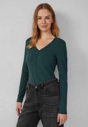 Groene geribbelde longsleeve top met een V-hals en knoopsluiting, gecombineerd met donkergrijze high-waisted jeans. Effen achtergrond.