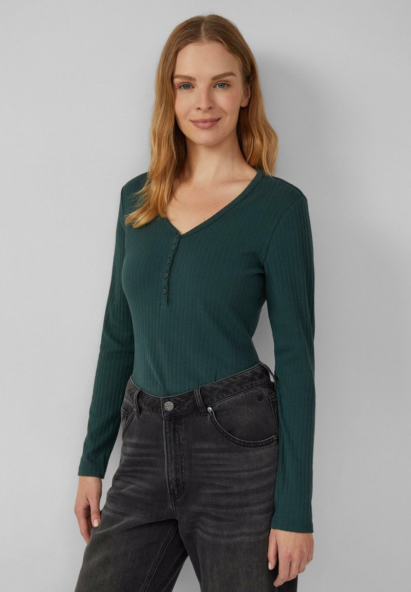 Grünes, geripptes Langarmoberteil mit V-Ausschnitt und Knopfverschluss, kombiniert mit dunkelgrauen, hochgeschnittenen Jeans. Einheitlicher Hintergrund.