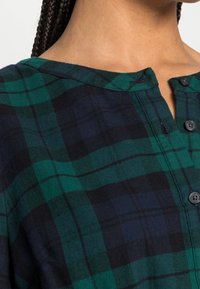 Chemise à carreaux verts et noirs avec un design à boutons, une texture douce et un col rond avec une petite patte de boutonnage et des boutons.