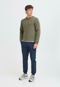 Olijfgroene gebreide trui met een ronde hals, gecombineerd met marineblauwe cargobroeken met zijzakken en elastische boorden. Grijze sneakers.