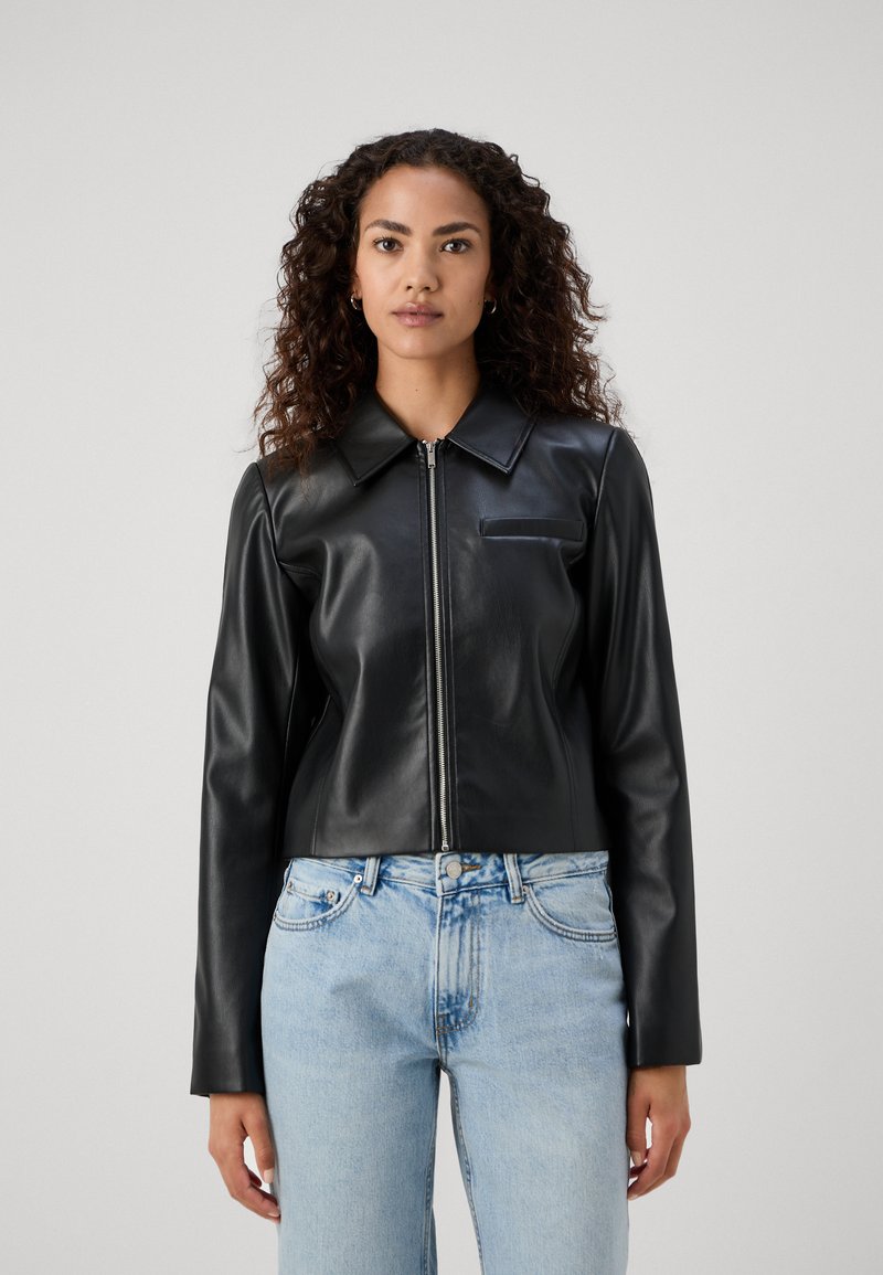 Giubbotto in pelle nera corto con zip frontale e colletto a punta. Presenta maniche lunghe e una piccola tasca sul petto. Indossato con jeans azzurri chiari.