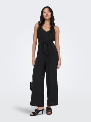 Kvinne står iført en svart ermeløs jumpsuit med midjebånd, svarte sandaler med hæl, og holder en liten svart håndveske.
