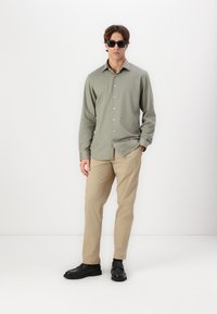 Chemise boutonnée vert olive clair, pantalon beige et chaussures noires. Le modèle porte des lunettes de soleil sombres et se tient devant un fond blanc uni.