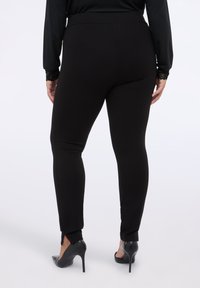 Černé fitted kalhoty s hladkou texturou a elastickým materiálem. Mají rozparky na kotnících a jsou kombinovány s černými vysokými podpatky.