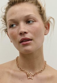 Ogrlica z zlatnim lancem koja ima privjesak u obliku zvijezde s teksturiranom površinom. Glatke karike spajaju privjesak s lancem.