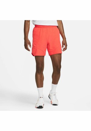 Mężczyzna w jasnokoralowych krótkich spodenkach sportowych, białych skarpetkach i białych trampkach stoi na tle jednolitego jasnego tła.