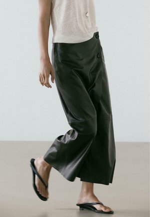 Personne portant un pantalon large en cuir noir, un haut léger sans manches et des sandales à talons noires, marchant sur un sol uni avec un fond clair.