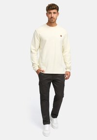 Helles gelbes Langarmhemd mit strukturiertem Muster, kombiniert mit dunklen Cargo-Hosen und weißen Sneakers. Kleines Logo auf der Brust.
