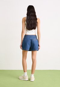 Τζιν σορτς με χαλαρή εφαρμογή, μπλε χρώμα, σχέδιο πέντε τσεπών και ραμμένο στρίφωμα, συνδυασμένα με λευκό αμάνικο μπλουζάκι και μπεζ αθλητικά παπούτσια.