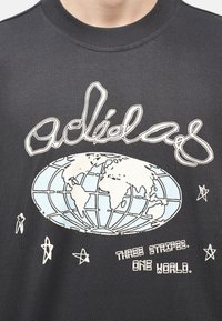 Σκούρο γκρι πουκάμισο με λευκό και γαλάζιο γήινο σφαιρικό σχέδιο, τη λέξη «adidas» από πάνω, αστέρια γύρω, και κείμενο που γράφει «THREE STRIPES. ONE WORLD.»