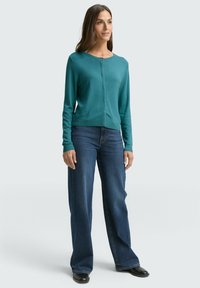 Zielony kardigan z okrągłym dekoltem, zapinany na guziki z przodu, z długimi rękawami i prążkowanym wykończeniem na dole. Połączony z niebieskimi szerokimi dżinsami i czarnymi butami.