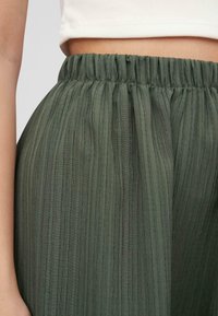 Grønne, plisserte bukser har elastisk liv, laget av lett stoff med en myk, teksturert overflate og vertikale striper.
