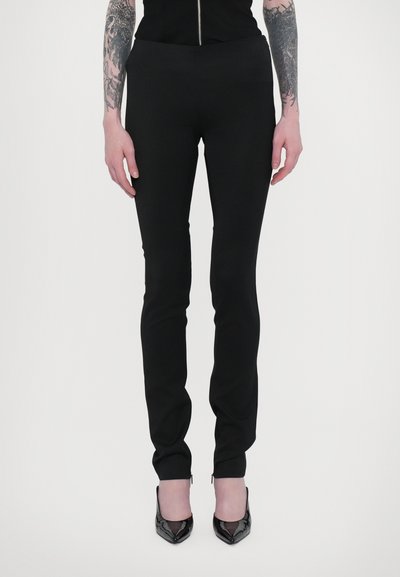 Black fitted pants with a smooth texture, featuring a slim design and small zip accents at the ankles. Worn with black pointed heels.