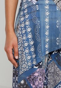 Main gauche reposant à côté d'une jupe en patchwork bleue et violette avec des motifs floraux et en paisley.