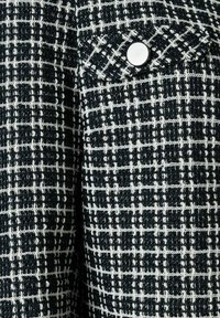 Schwarz-weißer Tweedstoff mit einem karierten Muster, ausgestattet mit einer kleinen Tasche und einem silbernen Knopfdetail. Strukturierten Oberfläche mit sichtbaren Nähten.