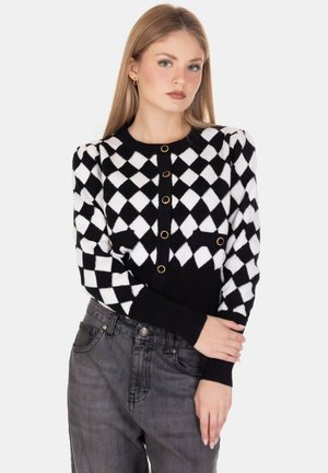 Cardigan a motivo argyle in bianco e nero con bottoni dorati, polsini a costine e una forma aderente, abbinato a jeans grigio scuro.