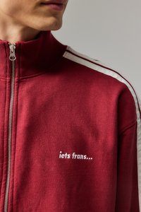 Punane tõmblukuga svetryhmed, millel on kõrge krae, valgete külgmiste triipudega ja tikandiga tekst "iets frans..." rinnal. Puuvillane materjal.