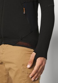 Crni dugosleeve majica s teksturiranim materijalom i narančastim patentnim zatvaračem, uparen s khaki hlačama koje imaju bočnu džepu. Mrežasti panel na rubu.