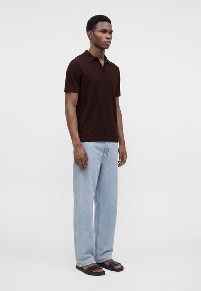 Jeune homme debout, portant un polo marron foncé à manches courtes, un jean bleu clair et des sandales noires sur un fond clair uni.
