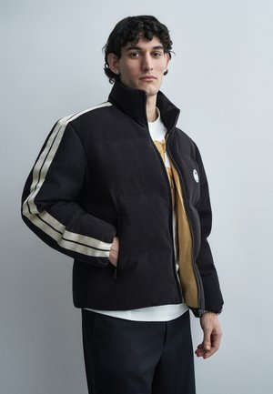 Νέος άνδρας που φοράει μαύρο μπουφάν με φερμουάρ και λευκές ρίγες στα μανίκια, με λευκό πουκάμισο από μέσα, στέκεται μπροστά σε απλό γκρι φόντο.